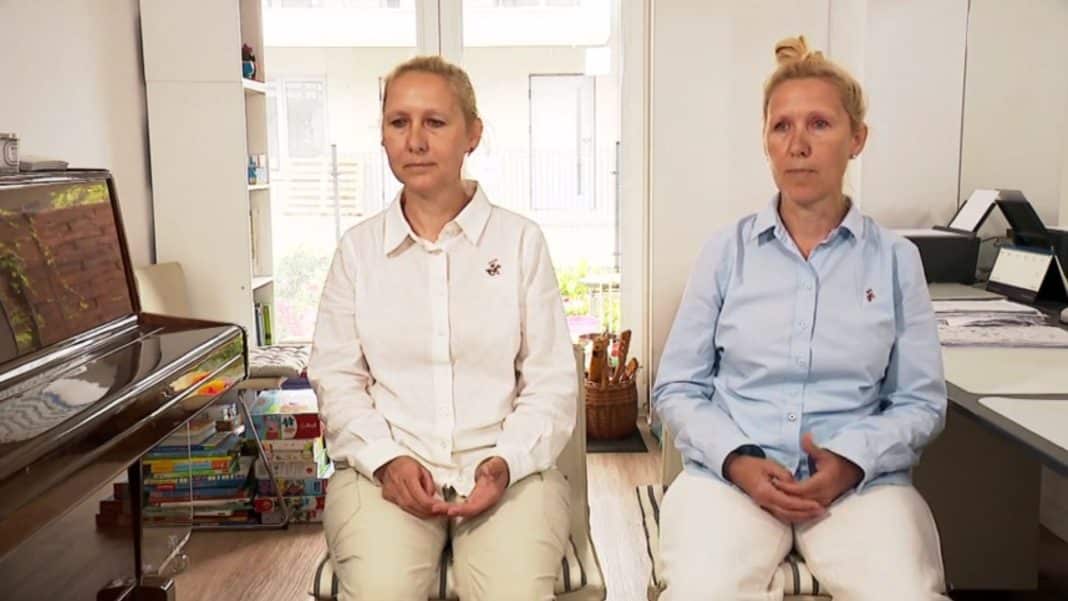 Dwie kobiety siedzą w biurze, obie mają blond włosy i noszą białe koszule, jedna z nich wygląda na zmartwioną.