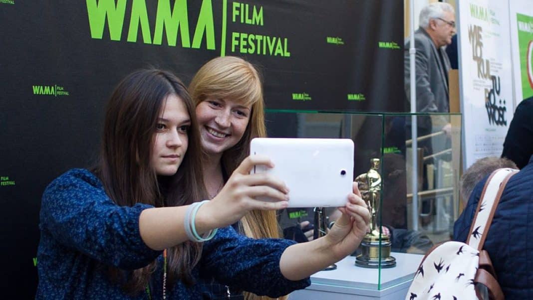 Dwie młode kobiety robią zdjęcie na tle plakatu WAMA Film Festival, jedna z nich uśmiecha się.