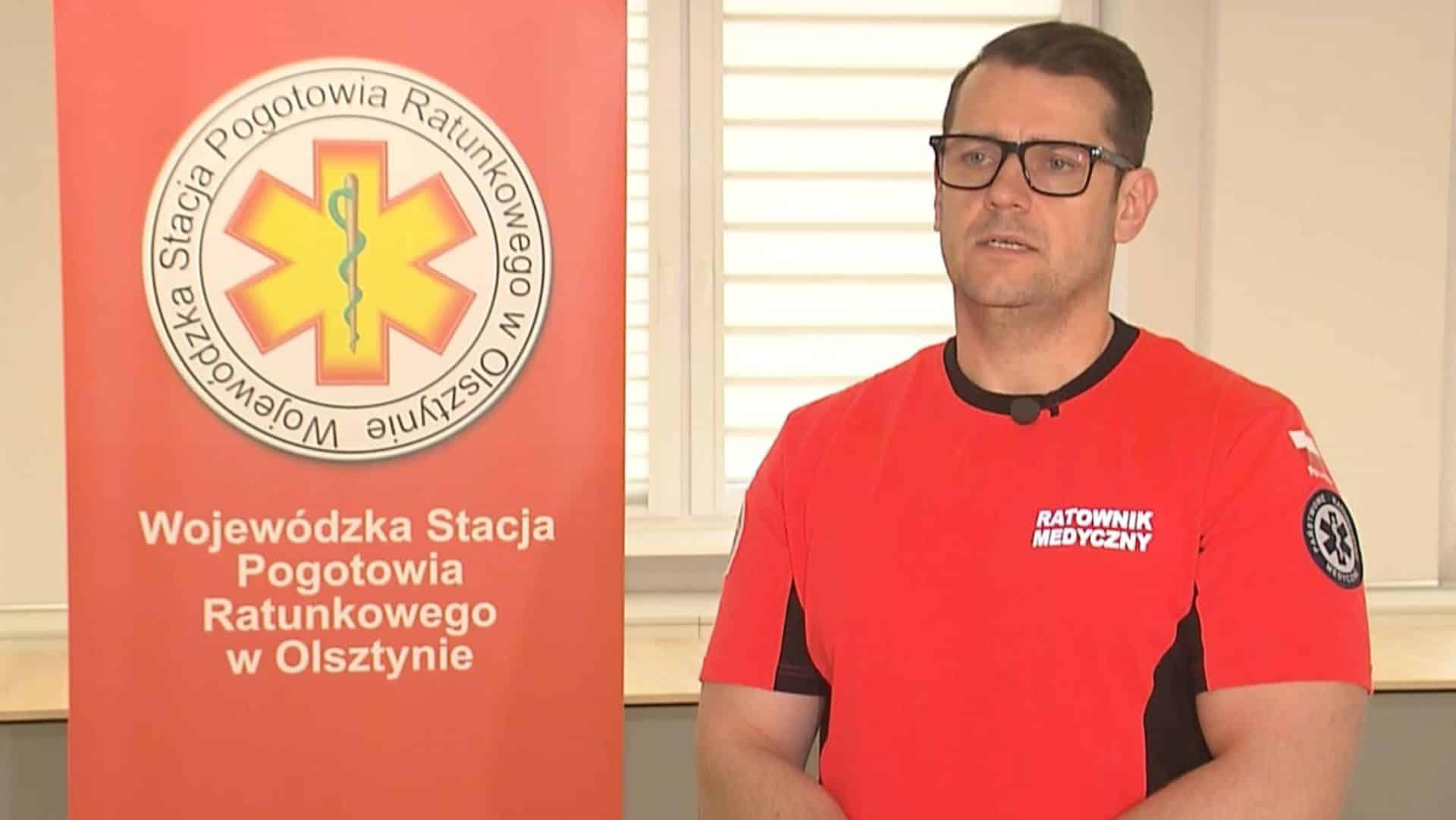 Mężczyzna w czerwonej koszulce z napisem 'Ratownik Medyczny' podczas prezentacji w pomieszczeniu, obok plakat z logo pogotowia ratunkowego.