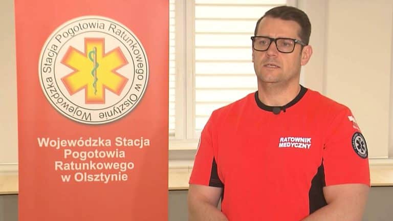 Mężczyzna w czerwonej koszulce z napisem 'Ratownik Medyczny' podczas prezentacji w pomieszczeniu, obok plakat z logo pogotowia ratunkowego.