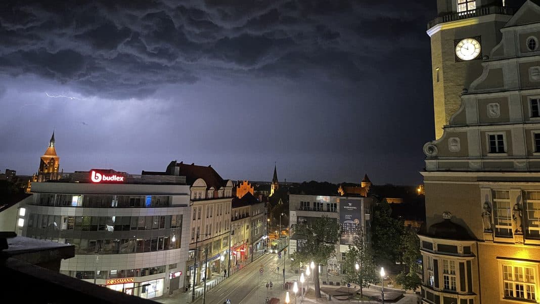 Burza z błyskawicami nad miastem nocą, ciemne chmury na niebie, oświetlone budynki i ulica.