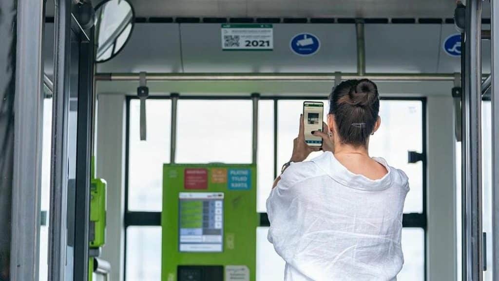 Kobieta skanuje kod QR na ekranie w autobusie w Olsztynie, widoczny jest zielony terminal biletowy.
