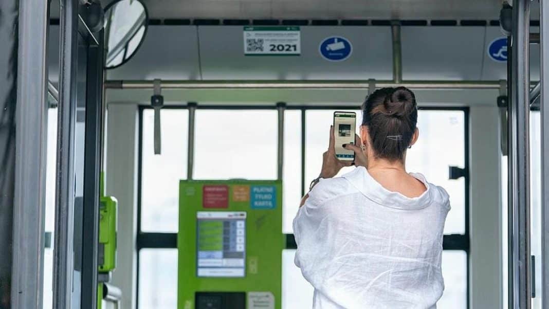 Kobieta skanuje kod QR na ekranie w autobusie w Olsztynie, widoczny jest zielony terminal biletowy.