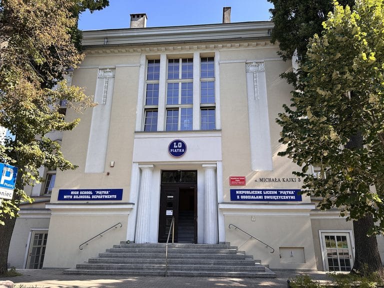Budynek liceum z dużymi oknami i tablicami informacyjnymi przy wejściu, otoczony drzewami.