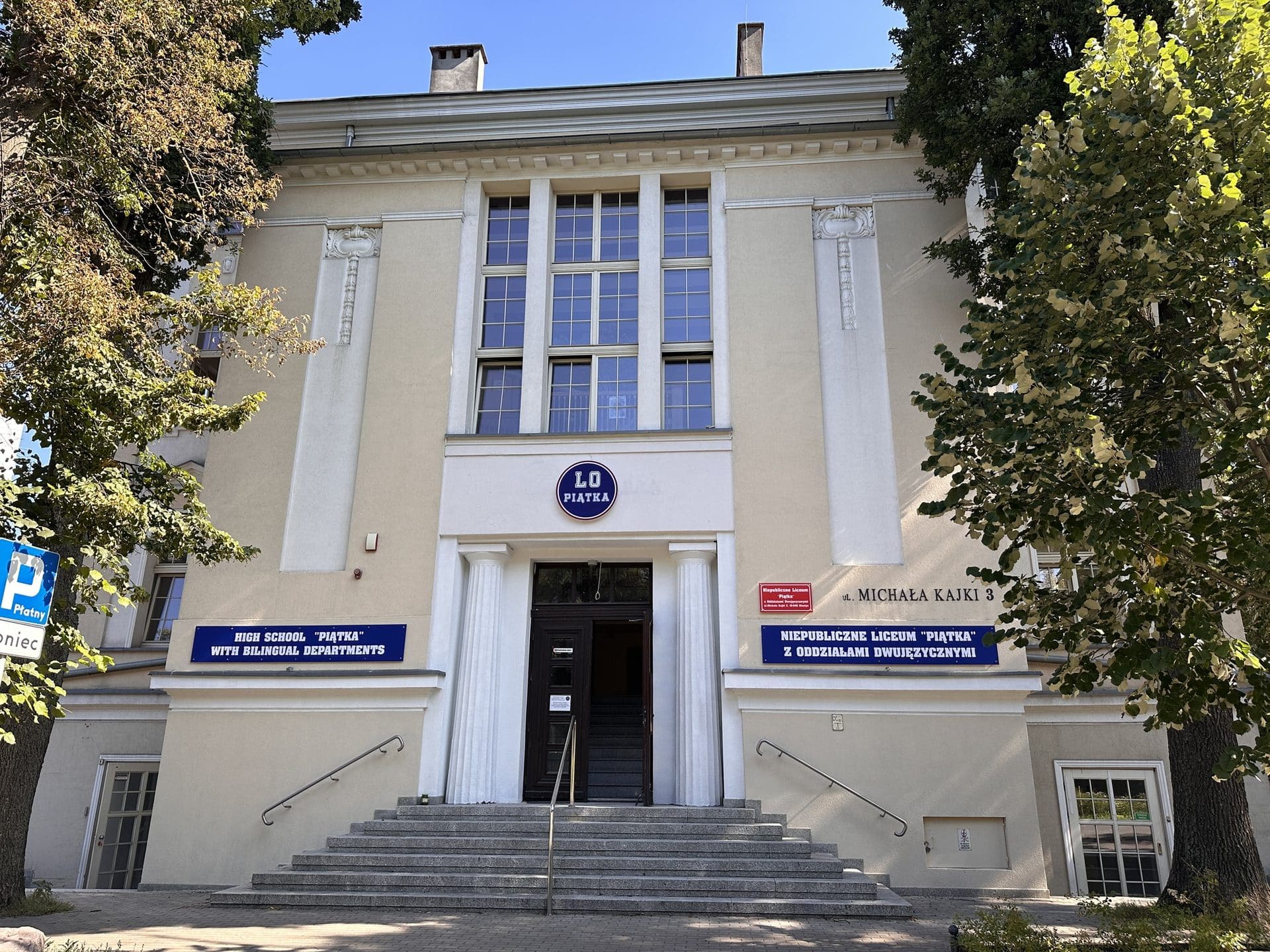 Budynek liceum z dużymi oknami i tablicami informacyjnymi przy wejściu, otoczony drzewami.