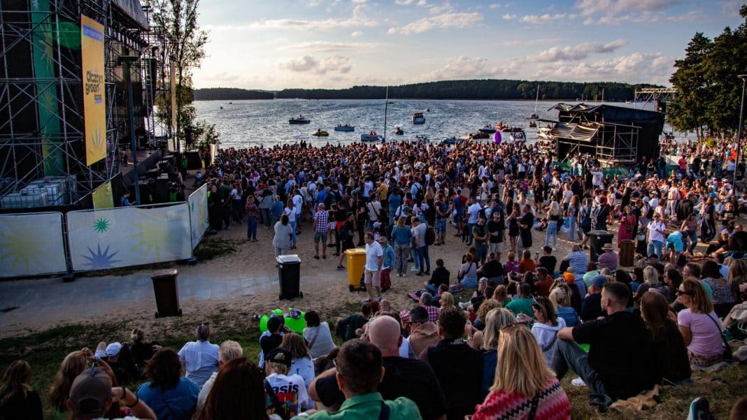 Tłum ludzi na festiwalowej plaży nad jeziorem podczas koncertu, scena i łodzie w tle.