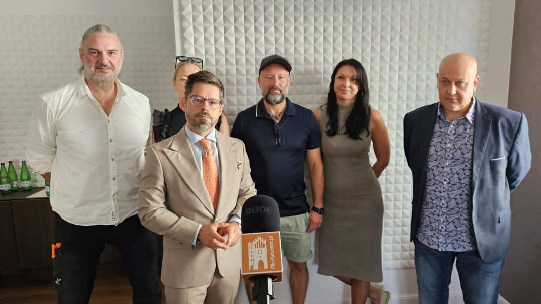 Grupa ludzi na konferencji prasowej, w tle ściana z paneli akustycznych.