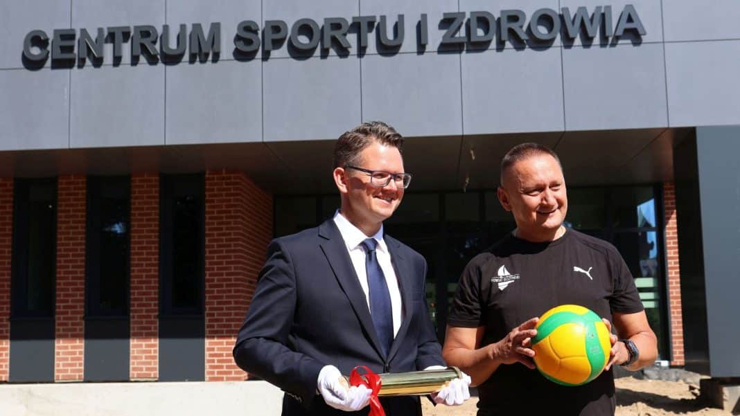 Dwa mężczyźni stoją przed nowoczesnym budynkiem z napisem 'Centrum Sportu i Zdrowia', jeden trzyma piłkę, drugi puchar.