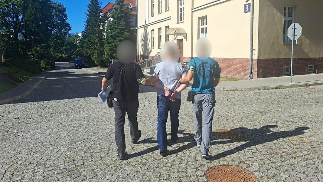 Mężczyzna w rękach policjantów na ulicy, z tyłu budynek i drzewa.