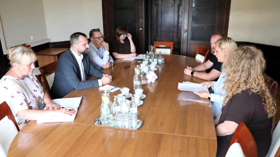 Grupa osób siedzi przy stole w sali konferencyjnej, rozmawiając podczas spotkania.
