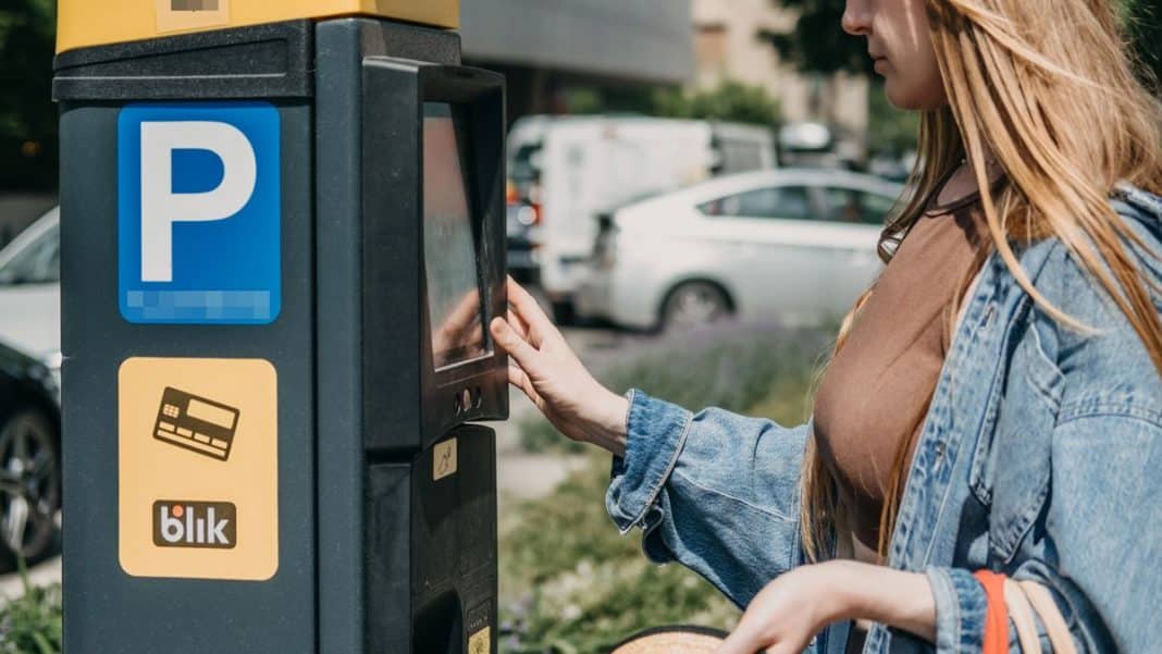 Kobieta korzysta z parkomatu w Olsztynie, w tle samochody na parkingu.