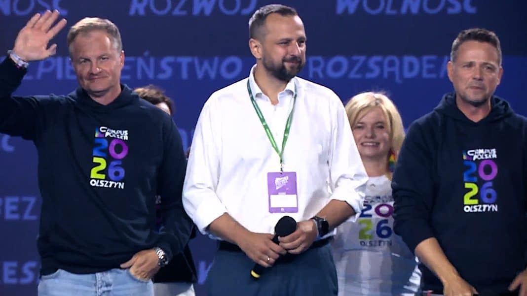 Grupa ludzi na scenie podczas wydarzenia, w tym mężczyzna w białej koszuli i dwaj mężczyźni w czarnych bluzach z logo wydarzenia.