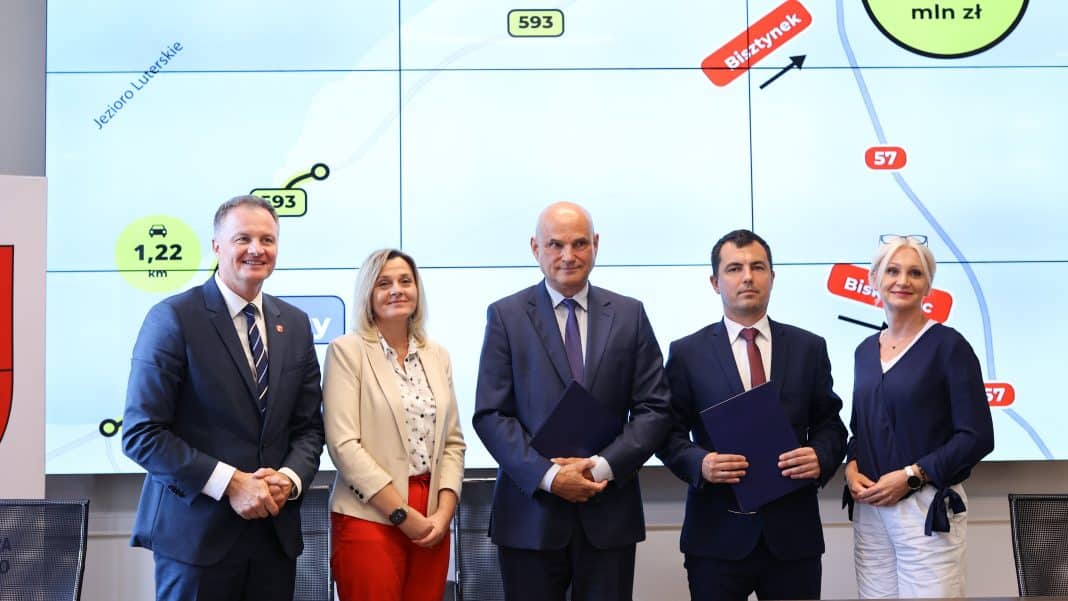 Grupa pięciu osób na tle dużego ekranu z mapą Warmii i Mazur, prezentujących plany inwestycji drogowych.