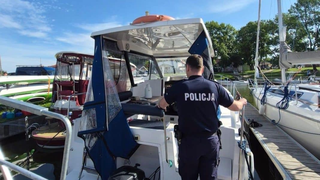 Policjant na pokładzie łodzi w porcie na Mazurach, wokół inne jachty i łodzie, słoneczna pogoda.