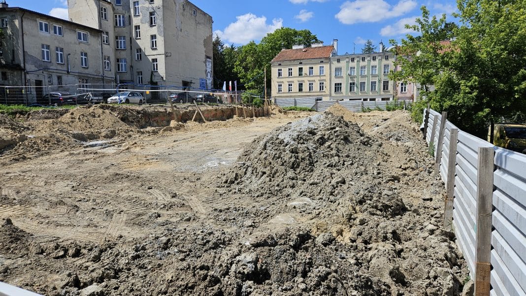 Plac budowy z wykopami i stosami ziemi, w tle stare kamienice i drzewa.