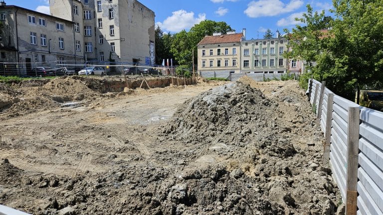 Plac budowy z wykopami i stosami ziemi, w tle stare kamienice i drzewa.