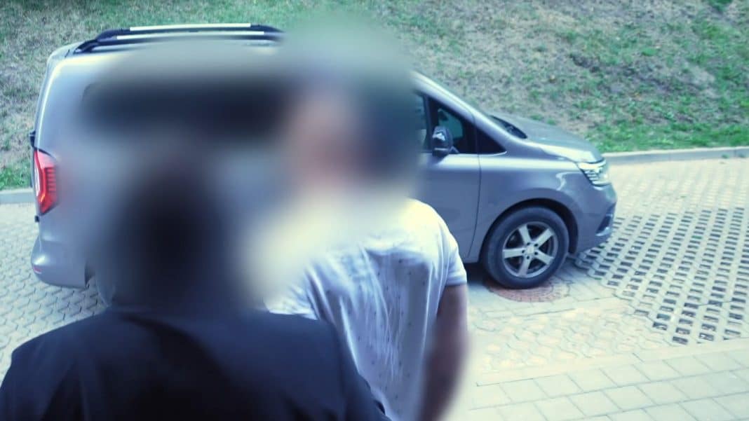 Mężczyzna w białej koszuli odwrócony plecami, stojący obok szarego samochodu na parkingu.