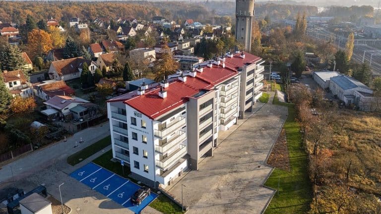 Widok na nowoczesny wielorodzinny budynek w Olsztynie z parkingiem i otaczającą zielenią.