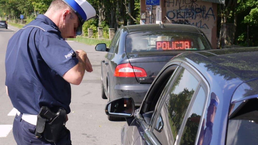 Policjant sprawdza dokumenty kierowcy podczas kontroli na drodze, w tle samochody i graffiti na ścianie.