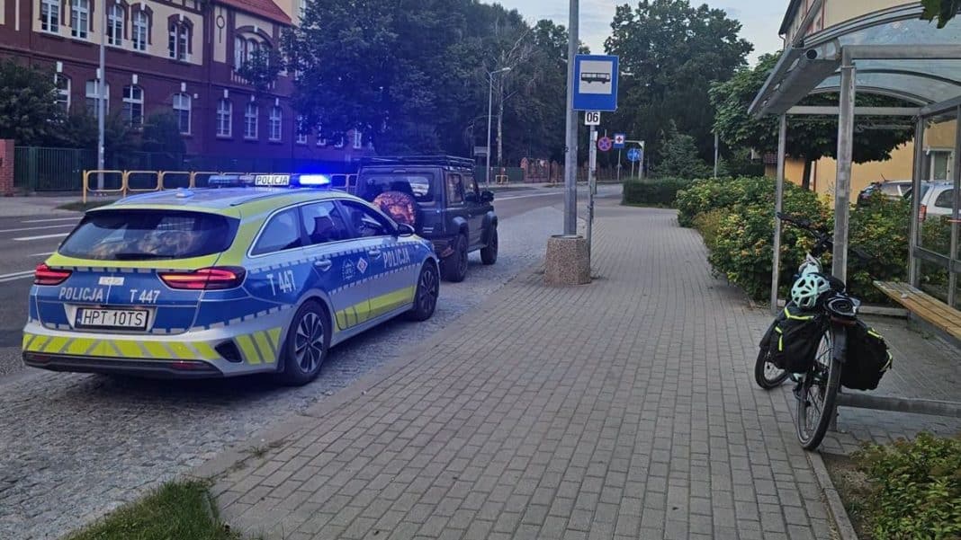 Na zdjęciu policyjny samochód na ulicy w Giżycku, obok rower z kaskiem i plecakiem, w tle budynki i drzewo.