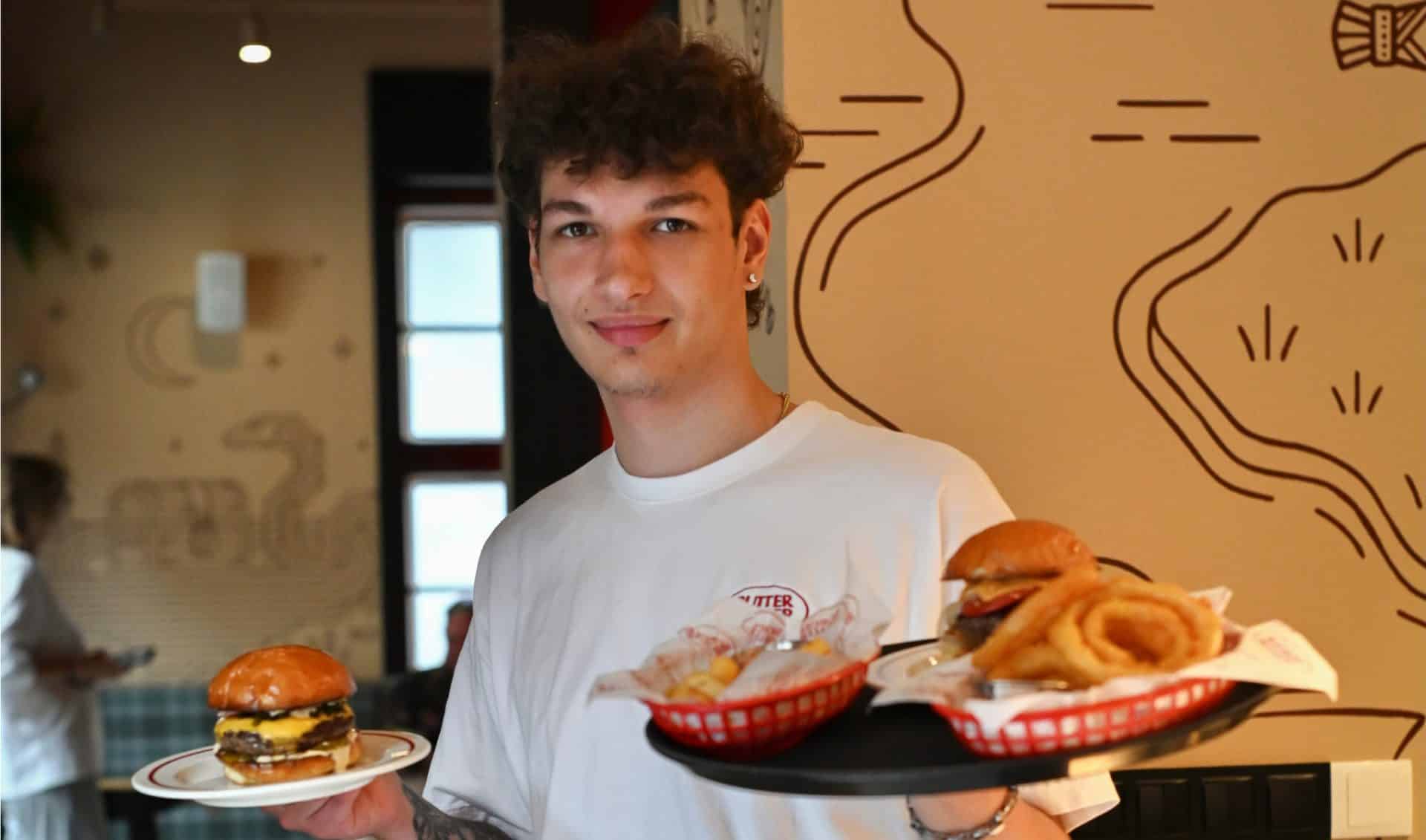 Mężczyzna trzyma dwa talerze z burgerami, w tle wnętrze restauracji z dekoracją na ścianie.