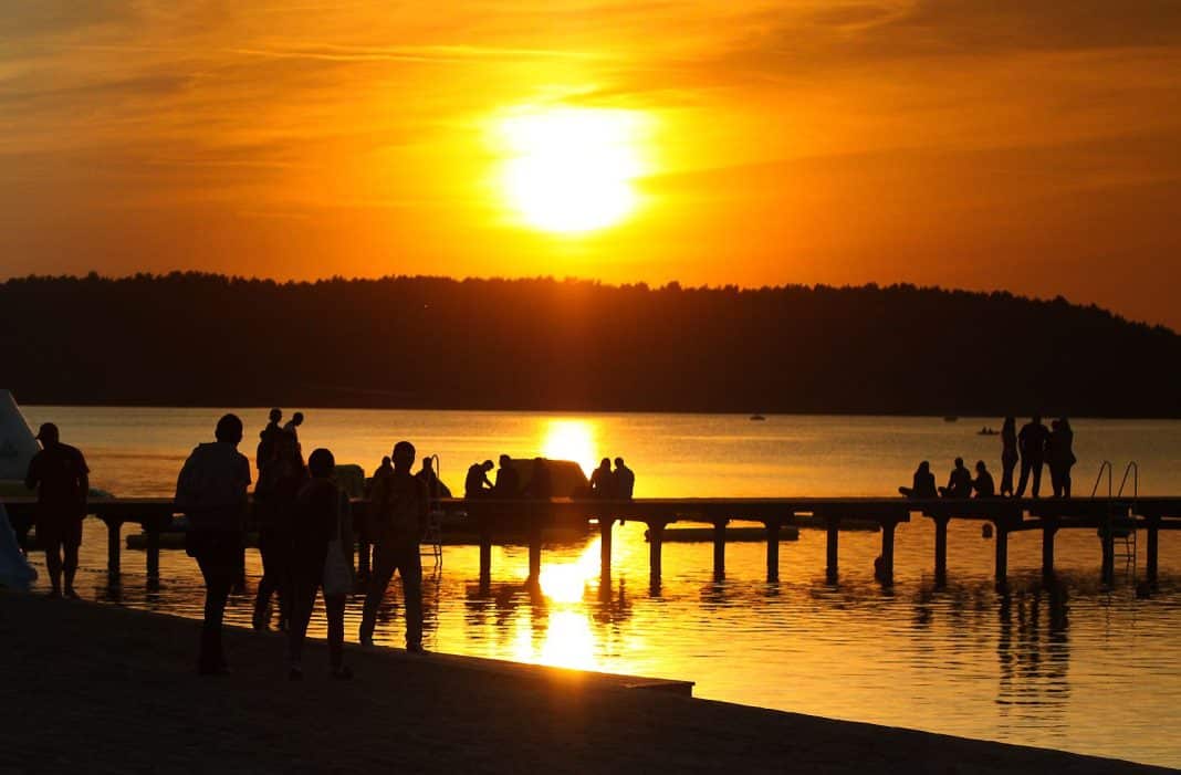 Grupa ludzi na plaży podczas zachodu słońca, widoczny księżyc przesłaniający część tarczy słonecznej.