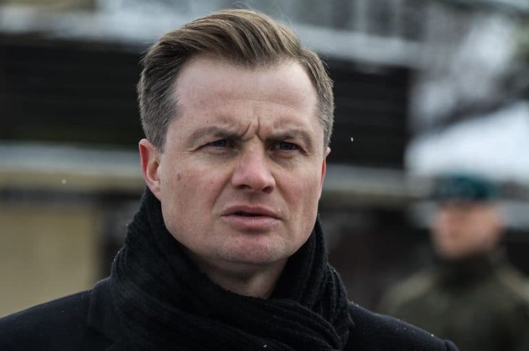 Mężczyzna na zewnątrz, ubrany w czarną kurtkę i szalik, z poważnym wyrazem twarzy.