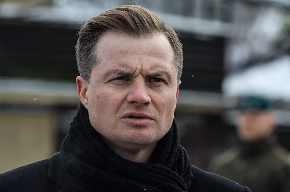 Mężczyzna na zewnątrz, ubrany w czarną kurtkę i szalik, z poważnym wyrazem twarzy.