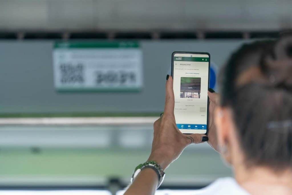 Kobieta trzyma telefon z kodem QR w autobusie miejskim, w tle tablica z rozkładem jazdy.