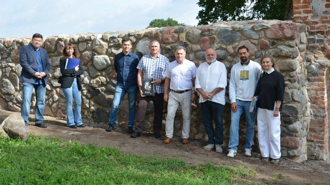 Grupa ludzi stoi przed odrestaurowanym fragmentem XIV-wiecznego muru z kamieni, przy bramie zamkowej.