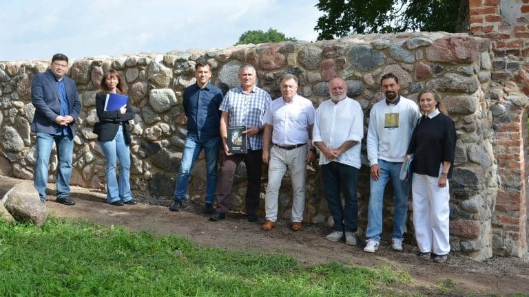 Grupa ludzi stoi przed odrestaurowanym fragmentem XIV-wiecznego muru z kamieni, przy bramie zamkowej.
