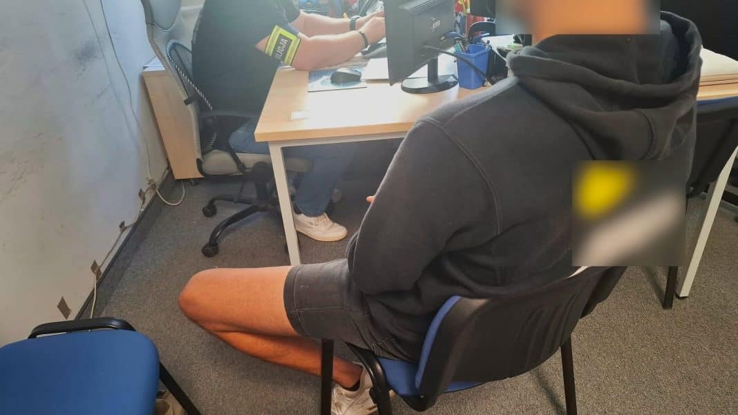 Mężczyzna siedzi na krześle w pomieszczeniu, z tyłu policjant przy biurku.