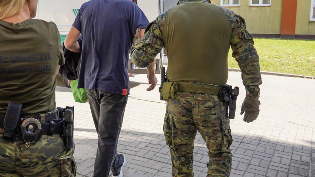 Funkcjonariusze Straży Granicznej odprowadzają cudzoziemca, w tle budynek mieszkalny.