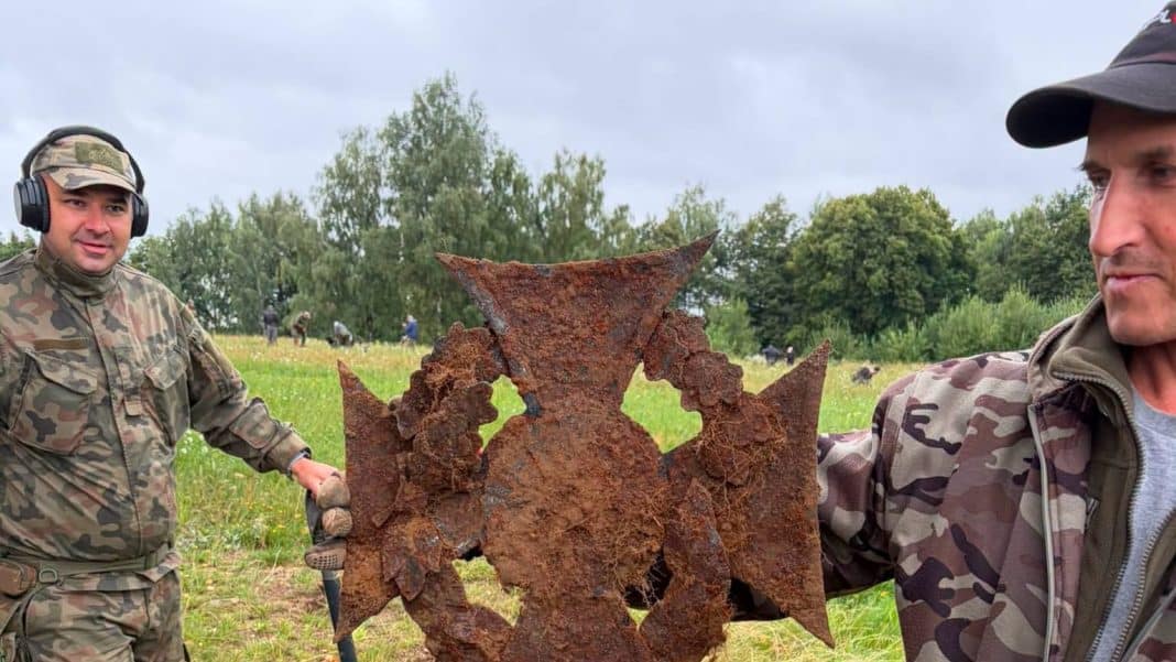 Dwa mężczyźni trzymają duży, stalowy krzyż harcerski na polu, w tle grupa ludzi i drzewa.