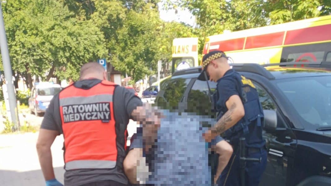 Ratownik medyczny i policjant udzielają pomocy mężczyźnie na ulicy w Olsztynie, w tle karetka pogotowia.