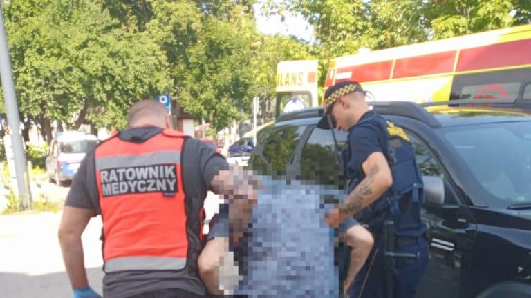 Ratownik medyczny i policjant udzielają pomocy mężczyźnie na ulicy w Olsztynie, w tle karetka pogotowia.