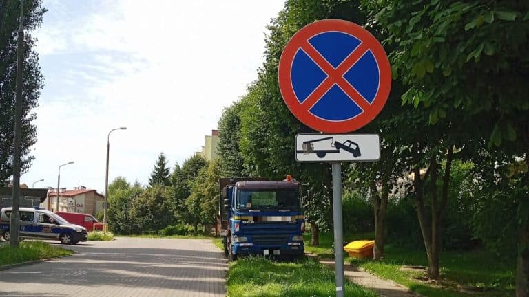 Ciężarowa auto-laweta zaparkowana przy zakazie, obok zielonych drzew i słupów oświetleniowych.