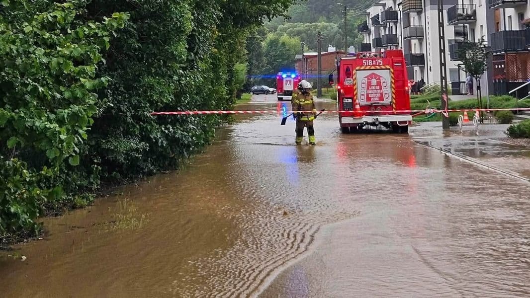 Strażacy na zalanej ulicy z wodą sięgającą do kolan, w tle bloki mieszkalne i drzewa.