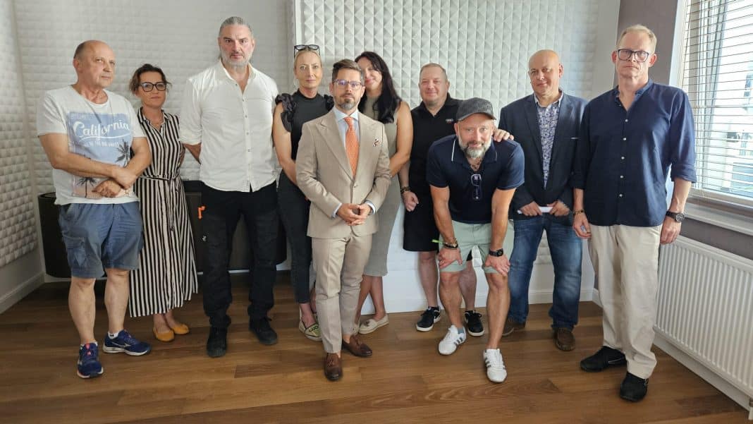 Grupa ludzi stojących w pomieszczeniu, niektórzy uśmiechają się, w tle okno z roletami.
