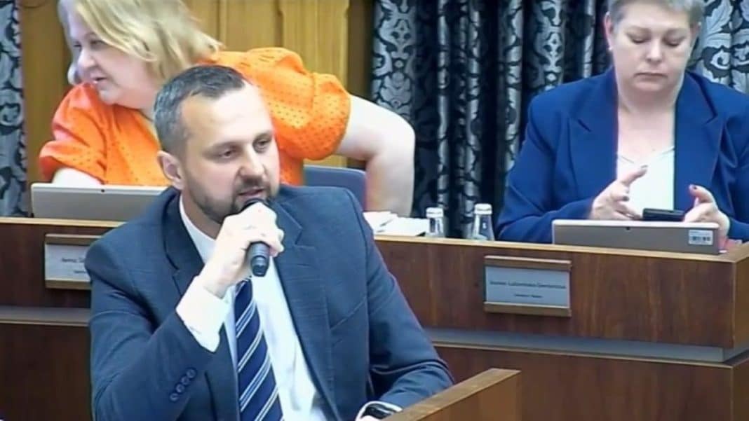 Osoby podczas sesji Rady Miasta Olsztyna, jedna z nich mówi do mikrofonu, w tle kobiety przy komputerach.