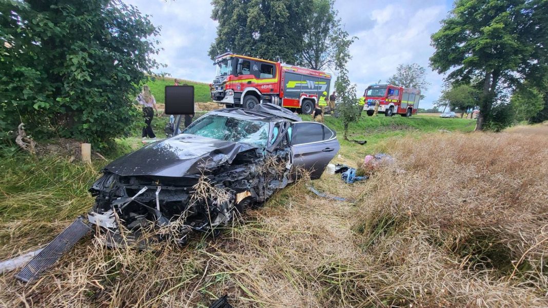 Rozbity samochód na poboczu drogi po wypadku, w tle straż pożarna i ratownicy.