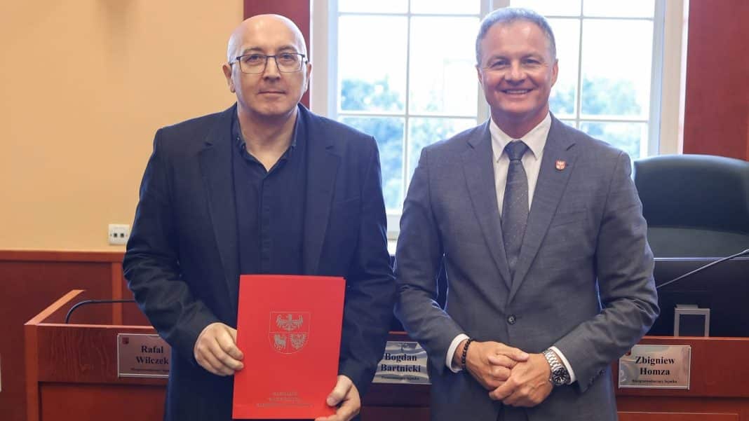 Dwóch mężczyzn w garniturach na tle okna, jeden z nich trzyma czerwony dokument, w tle tabliczki z nazwiskami.