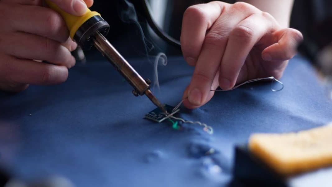 Osoba lutuje element elektroniczny za pomocą lutownicy z topnikiem, na stole znajduje się kabel i narzędzia.