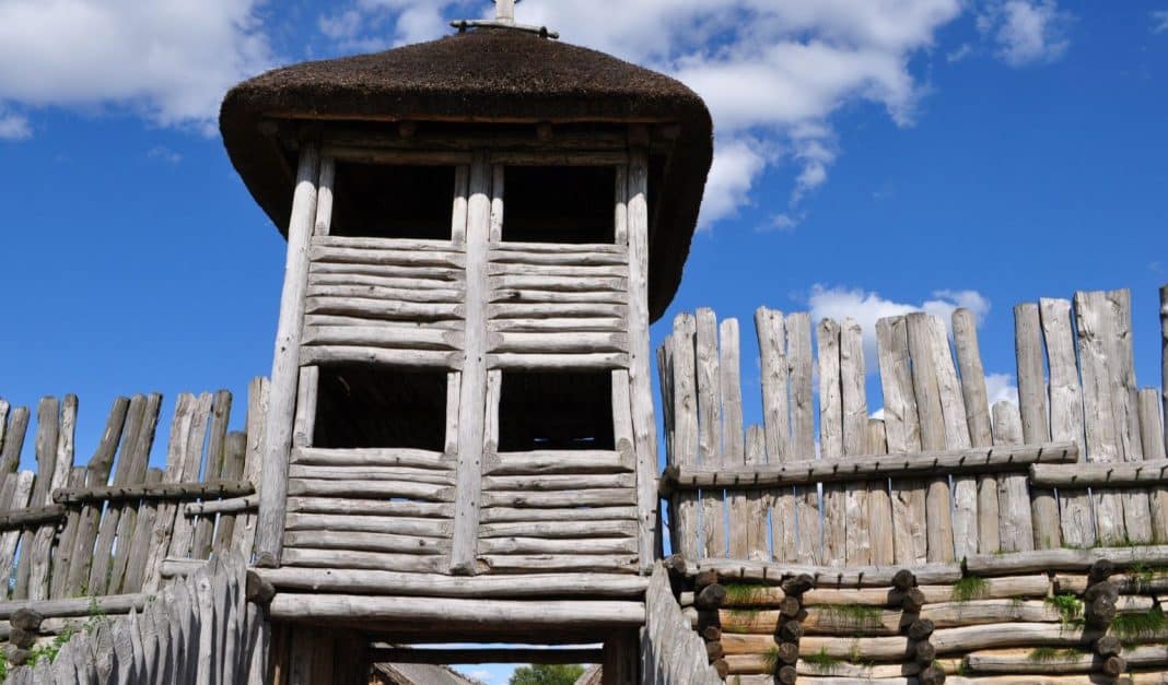 Drewniana wieża i palisada w skansenie archeologicznym w Biskupinie, pod niebieskim niebem.