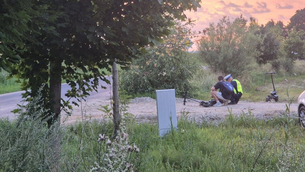 Policjant i młody chłopak przy hulajnodze na poboczu drogi, wieczorem, w otoczeniu drzew i zieleni.