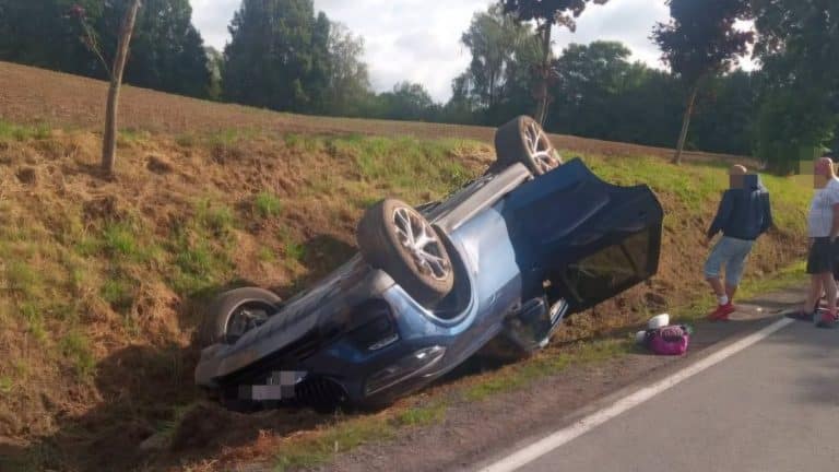 Rozbity samochód leży na boku przy drodze krajowej, wokół ludzie i służby ratunkowe.