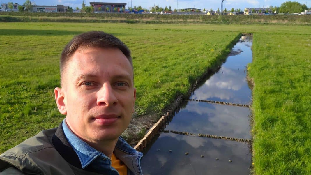 Przyrodnik na tle podmokłej łąki z gniazdami ptaków i pisklętami, w tle ulice i budynki.