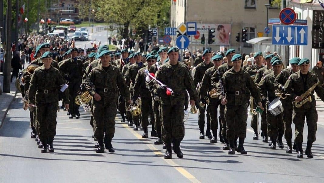 Uczestnicy wojskowego przemarszu w centrum miasta, w mundurach i z instrumentami muzycznymi.