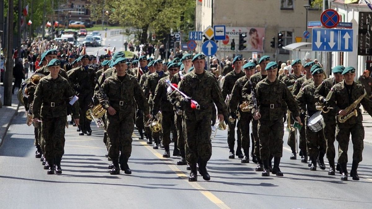 Uczestnicy wojskowego przemarszu w centrum miasta, w mundurach i z instrumentami muzycznymi.