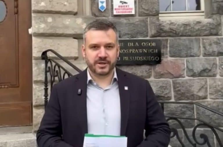 Mężczyzna w garniturze trzyma dokumenty przed budynkiem z tablicą informacyjną.